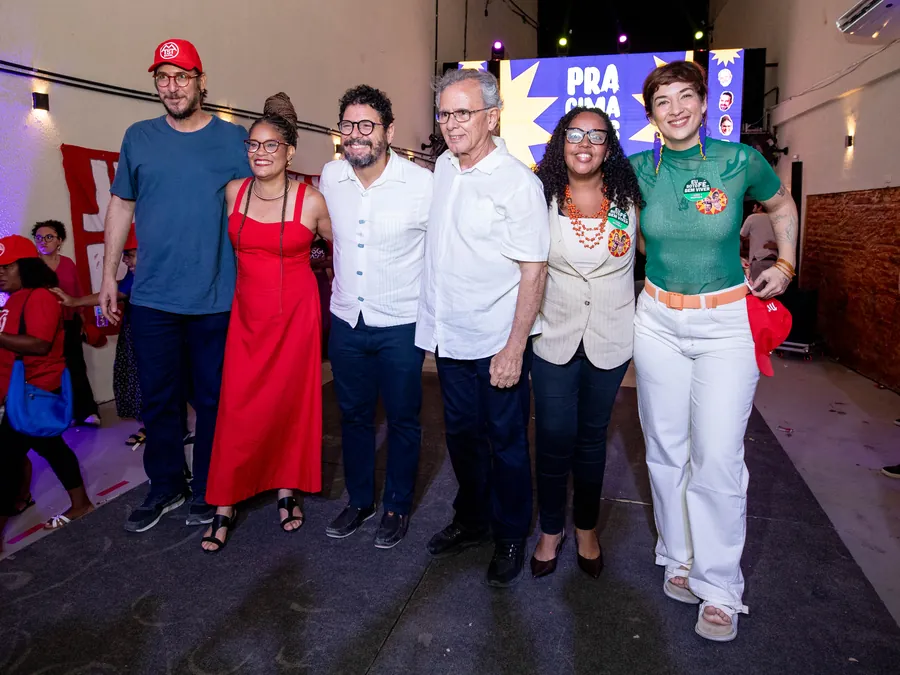 Bruna Albuquerque ao lado de aliadas e aliados da campanha em um encontro da federação, com bandeiras de movimentos populares ao fundo.