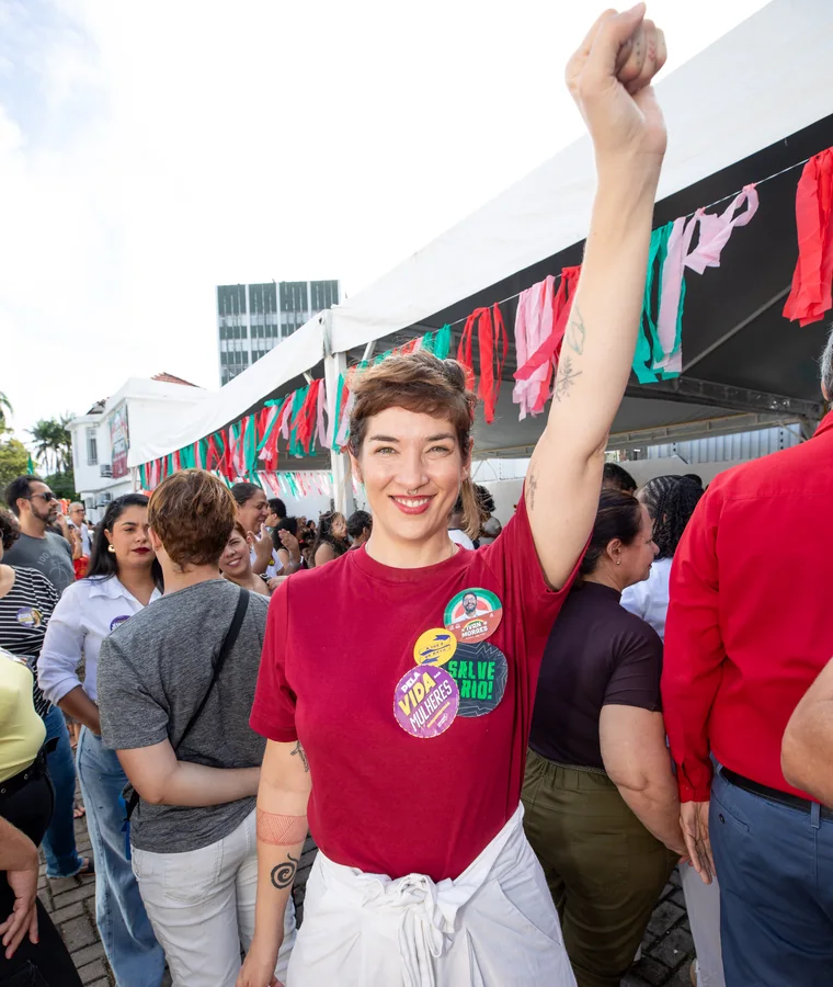 Bruna Albuquerque de punho erguido em um ato de rua, com broches na camiseta, cercada de gente.