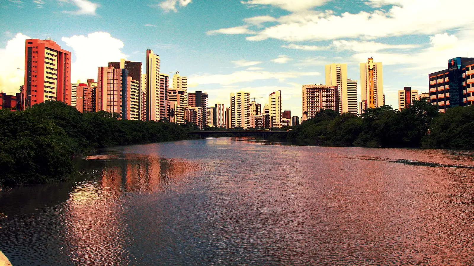 O rio Capibaribe atravessando o Recife ao fim da tarde, com os prédios da cidade ao fundo.